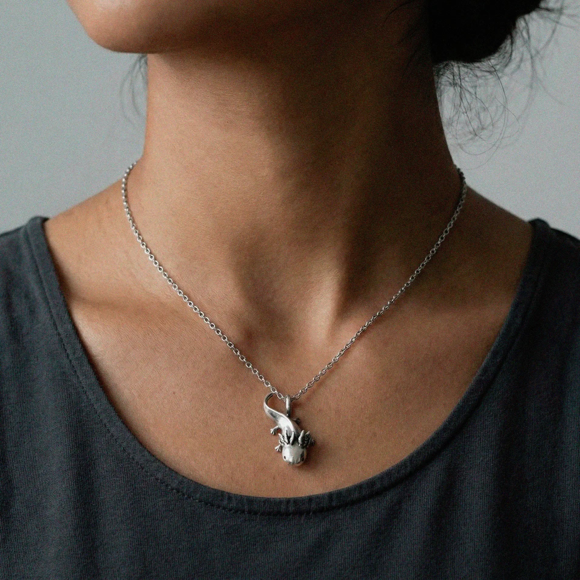 Person wearing a silver necklace with a small axolotl pendant against a neutral background
