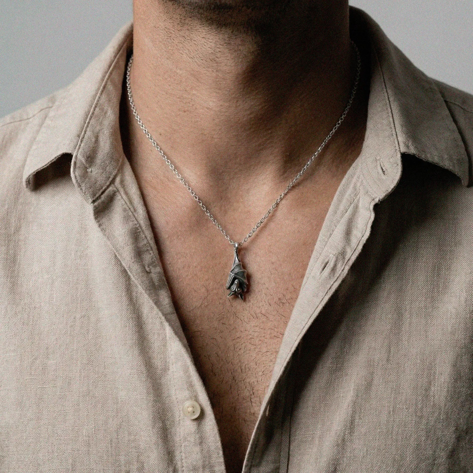 Man wearing a beige shirt with a necklace featuring a BAT pendant against a gray background