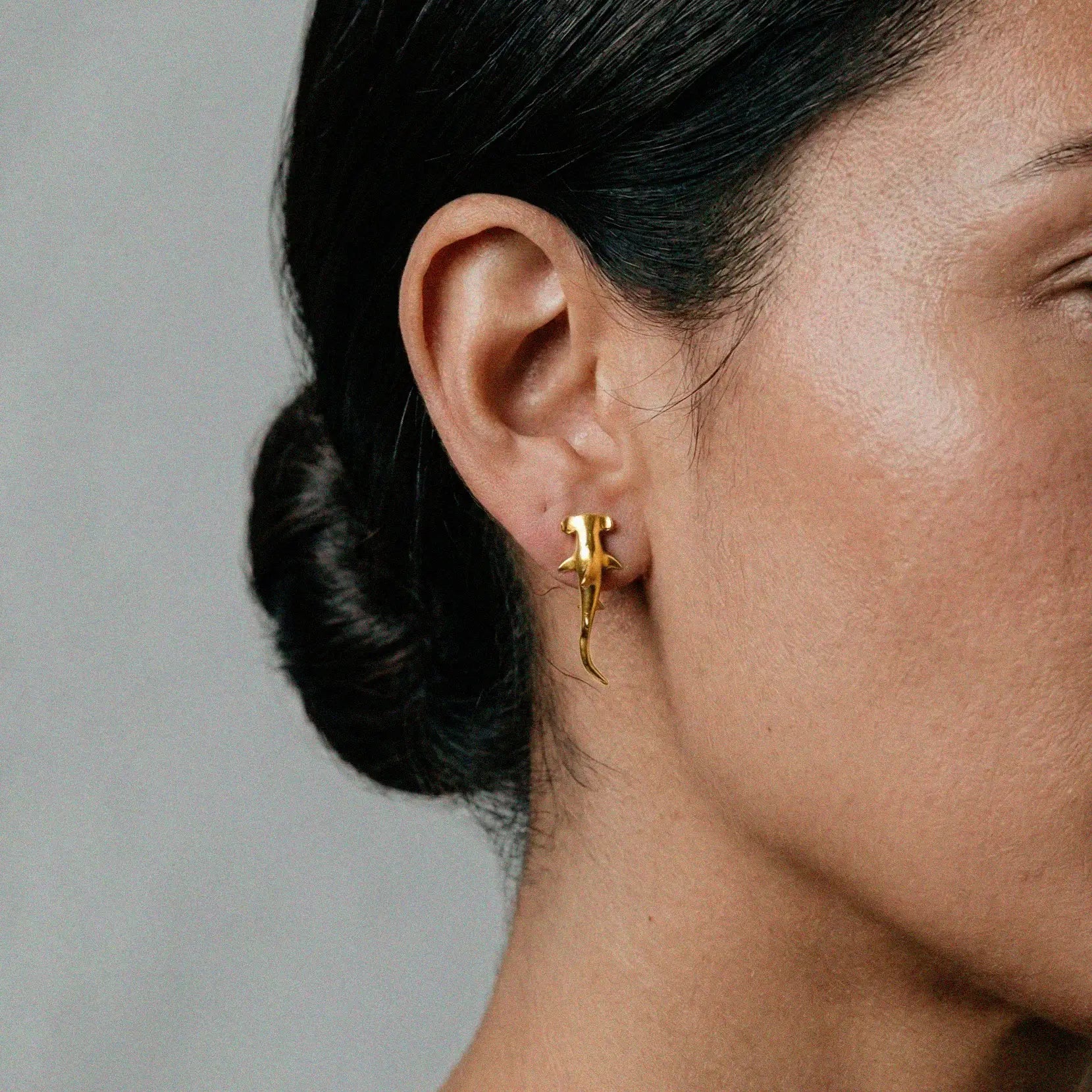 Close-up of a person wearing a gold  hammerhead-shark earring against a neutral background