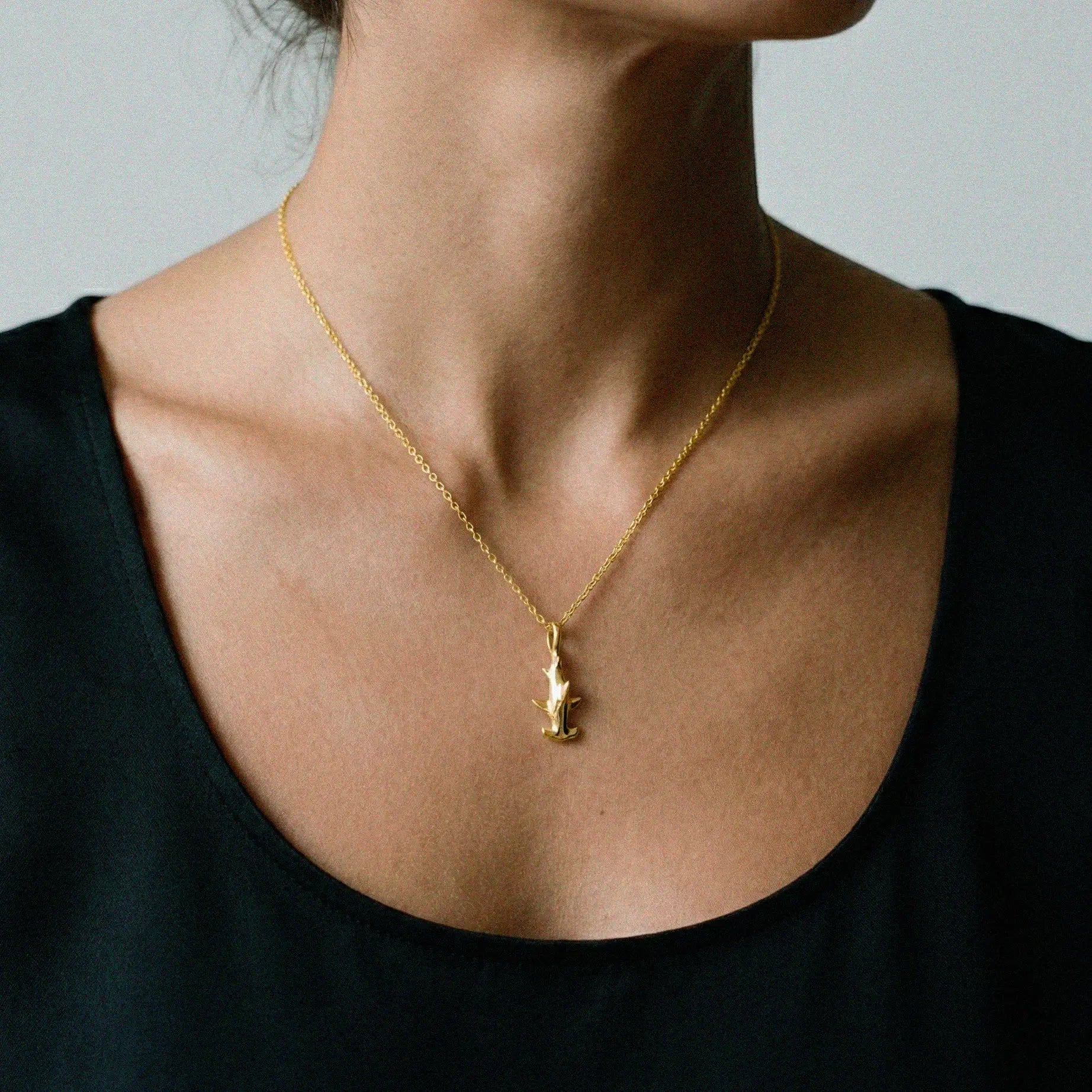 Gold necklace with a hammerhead-shark pendant on a person wearing a black top against a neutral background