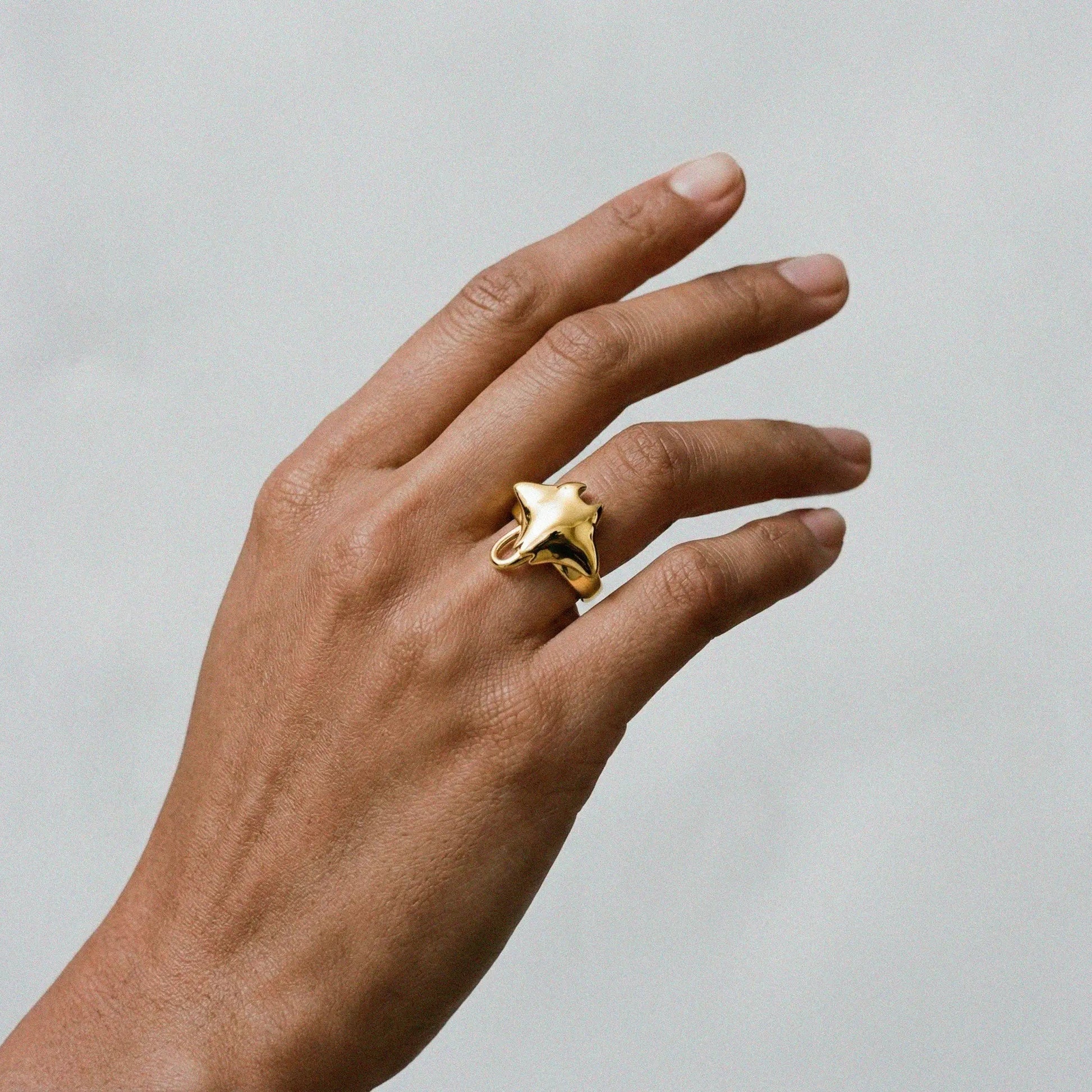 Hand wearing a gold ring with a manta ray design on a neutral background