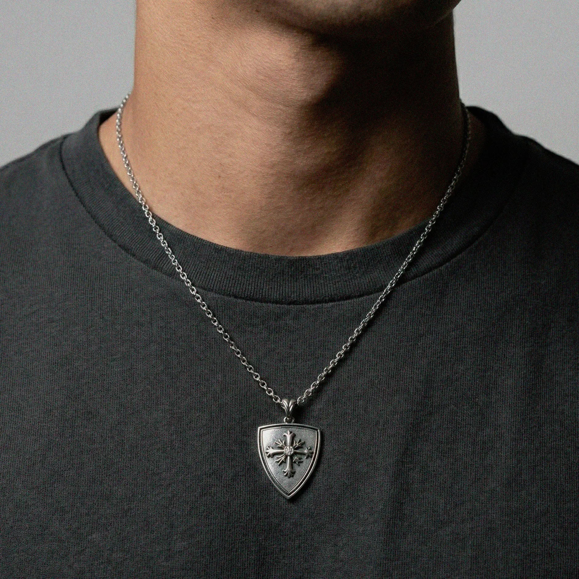 Person wearing a necklace with a shield-shaped pendant on a plain background
