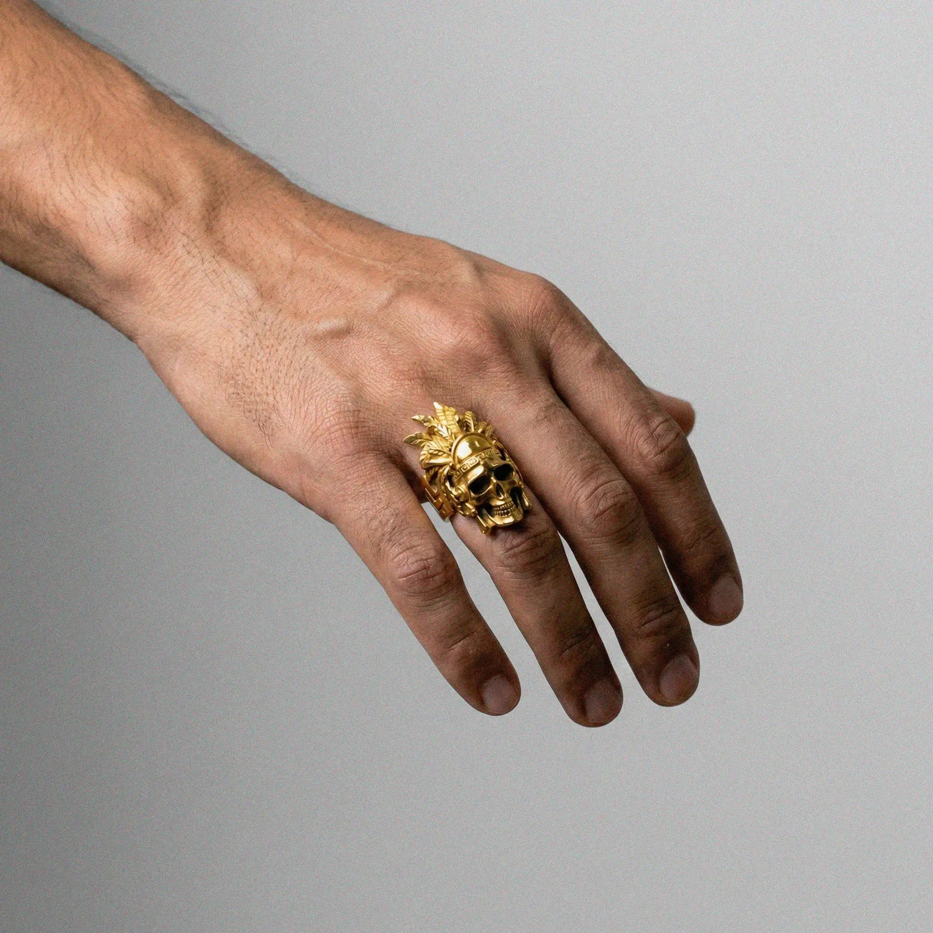 Hand wearing a gold ring with a skull with headdress on a gray background