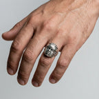 Hand wearing a silver ring with an olmec head design on a gray background