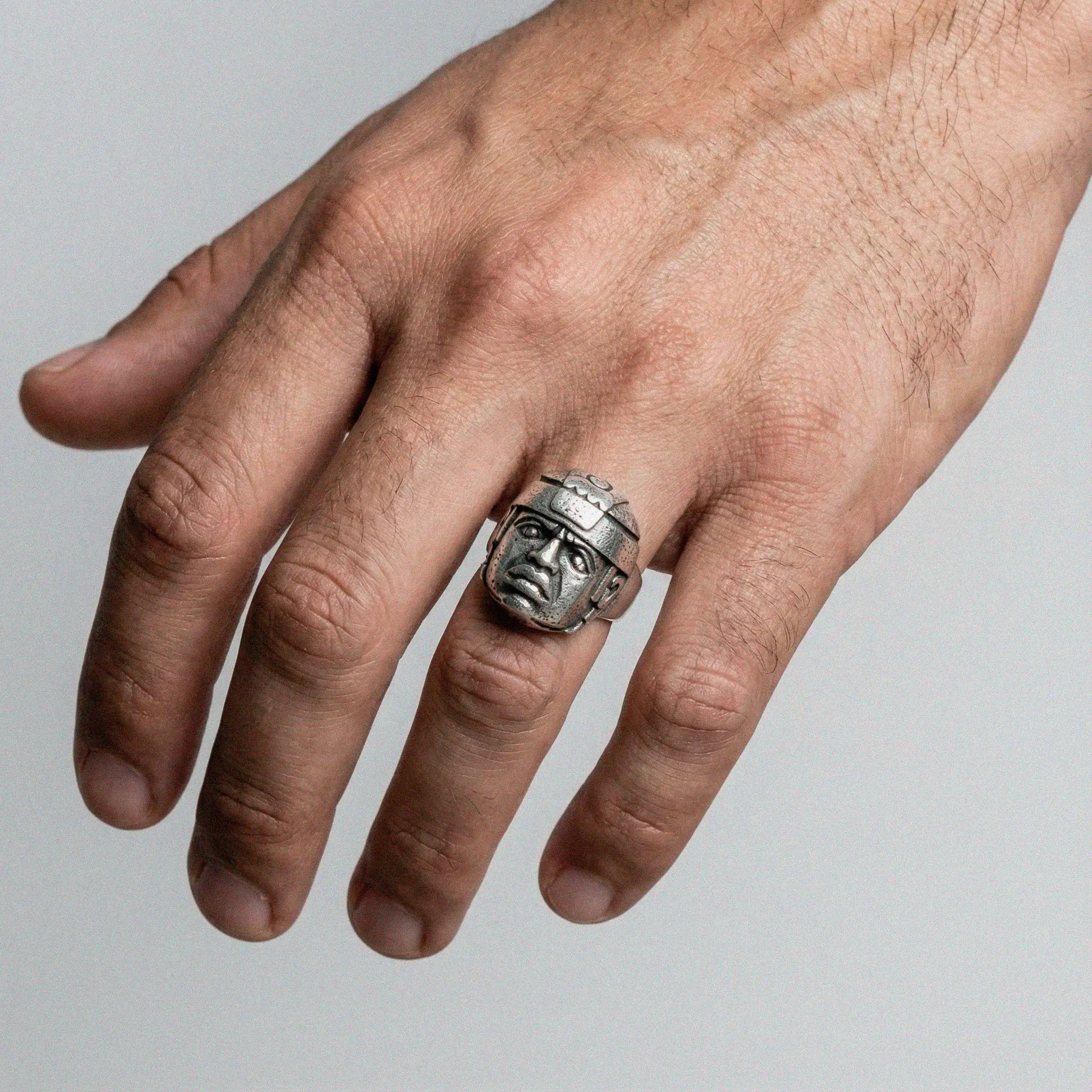 Hand wearing a silver ring with an olmec head design on a gray background