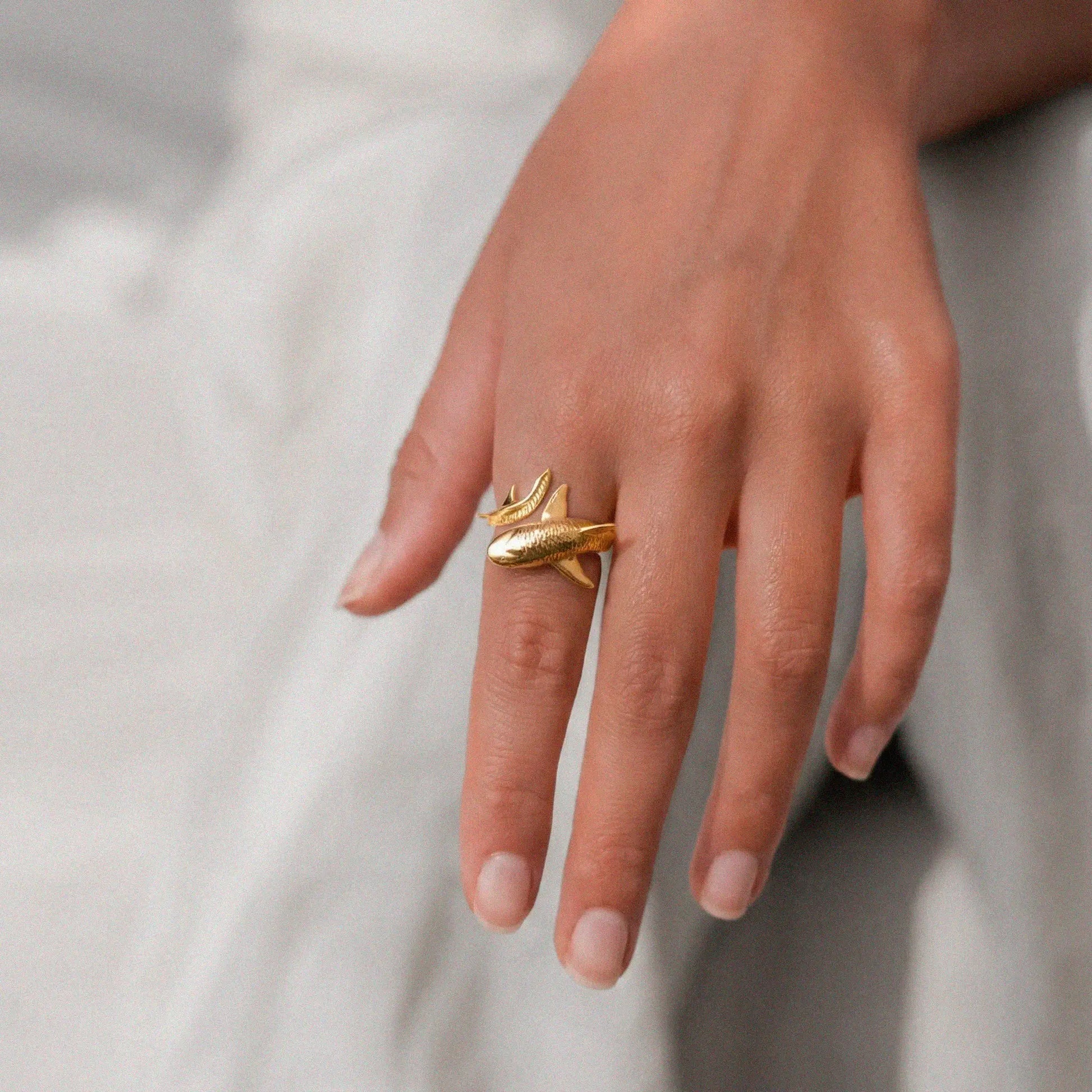Gold tiger-shark ring on a person's finger against a neutral background