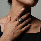 Close-up of a hand wearing a silver ring with tlaloc design, touching the neck against a neutral background.