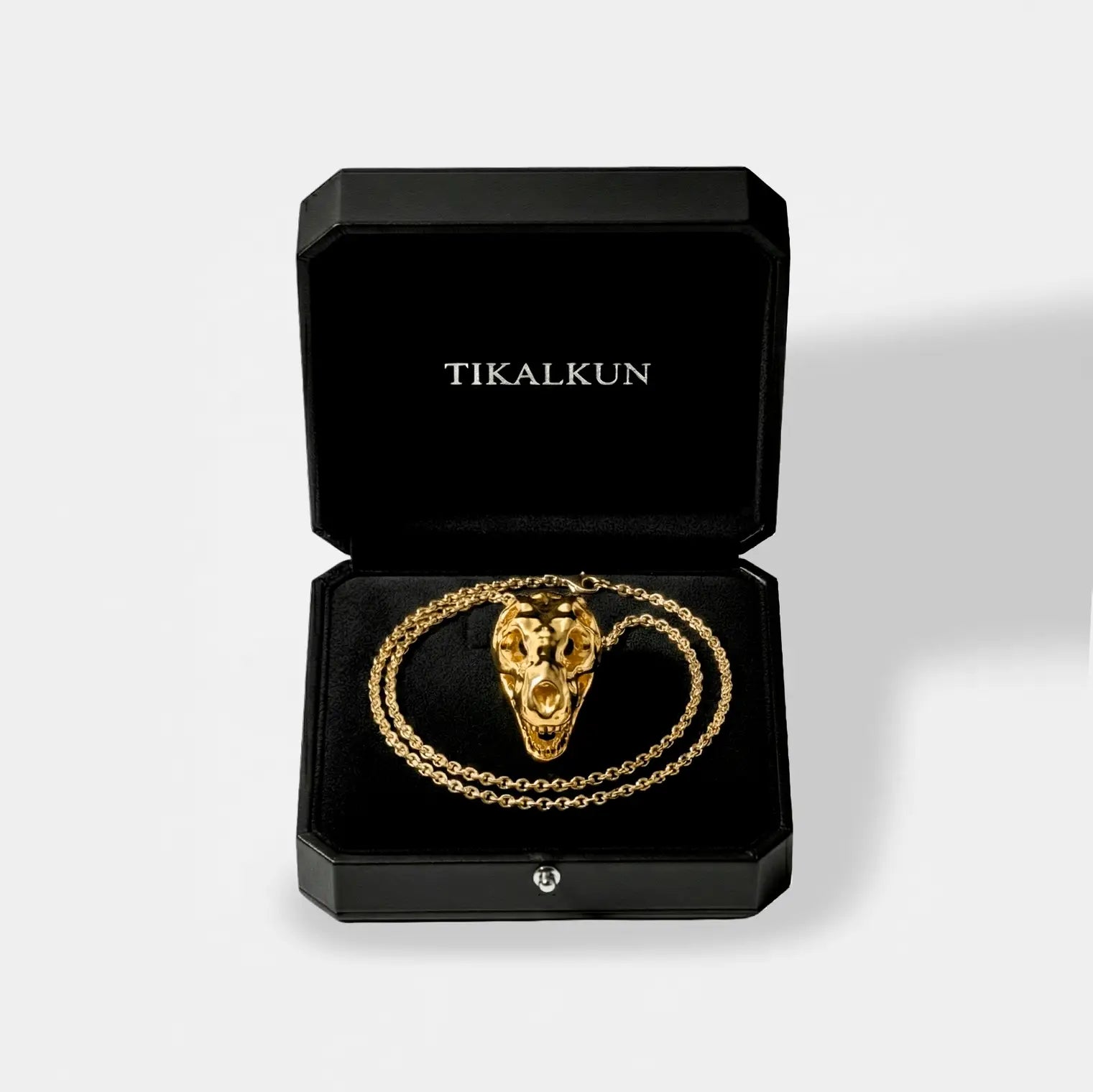 Gold necklace with t-rex skull design in a black Tikalkun box on a light gray background