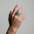 Hand wearing a silver venetian mask ring on a plain background