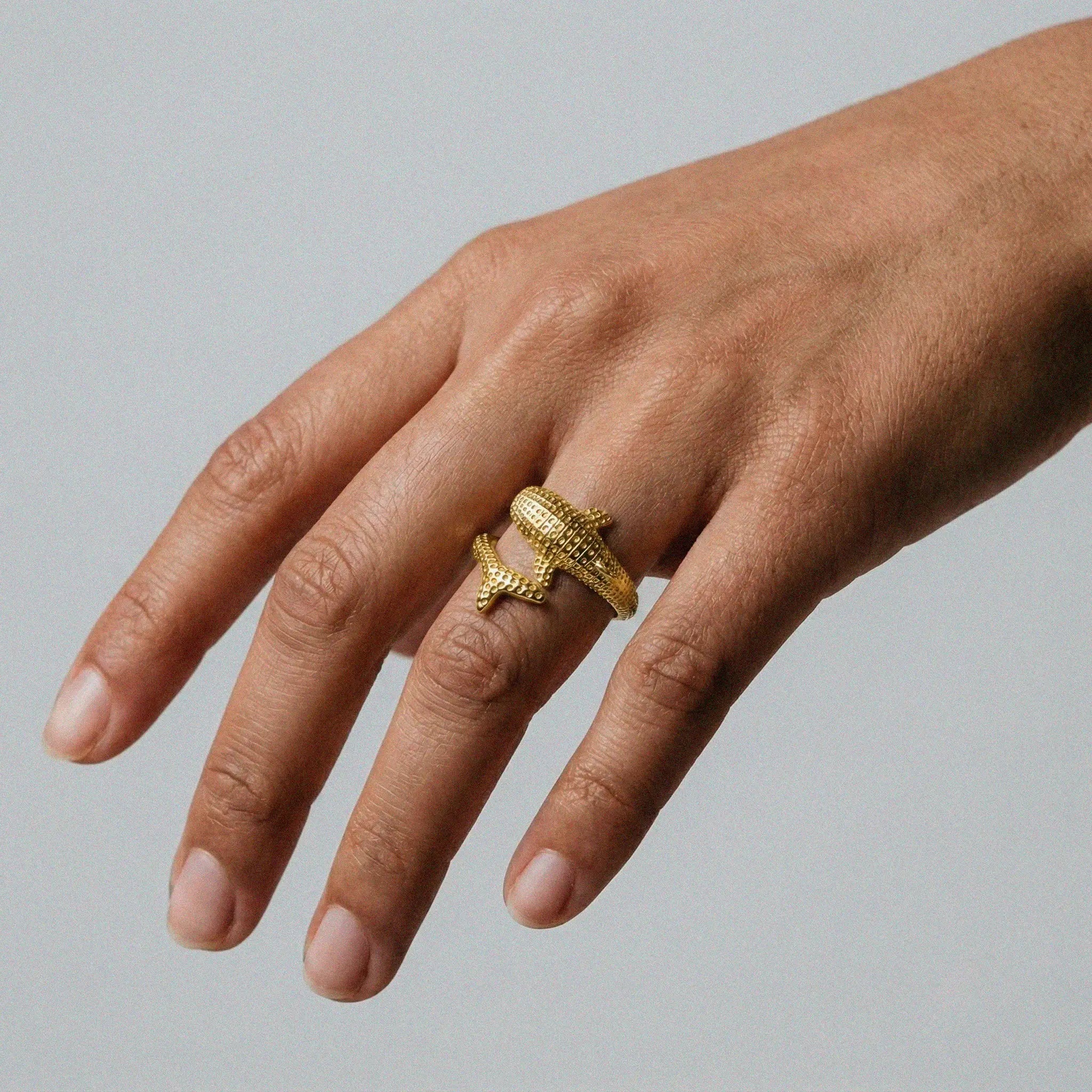 Hand wearing a gold whale-shark ring with a detailed design on a plain background