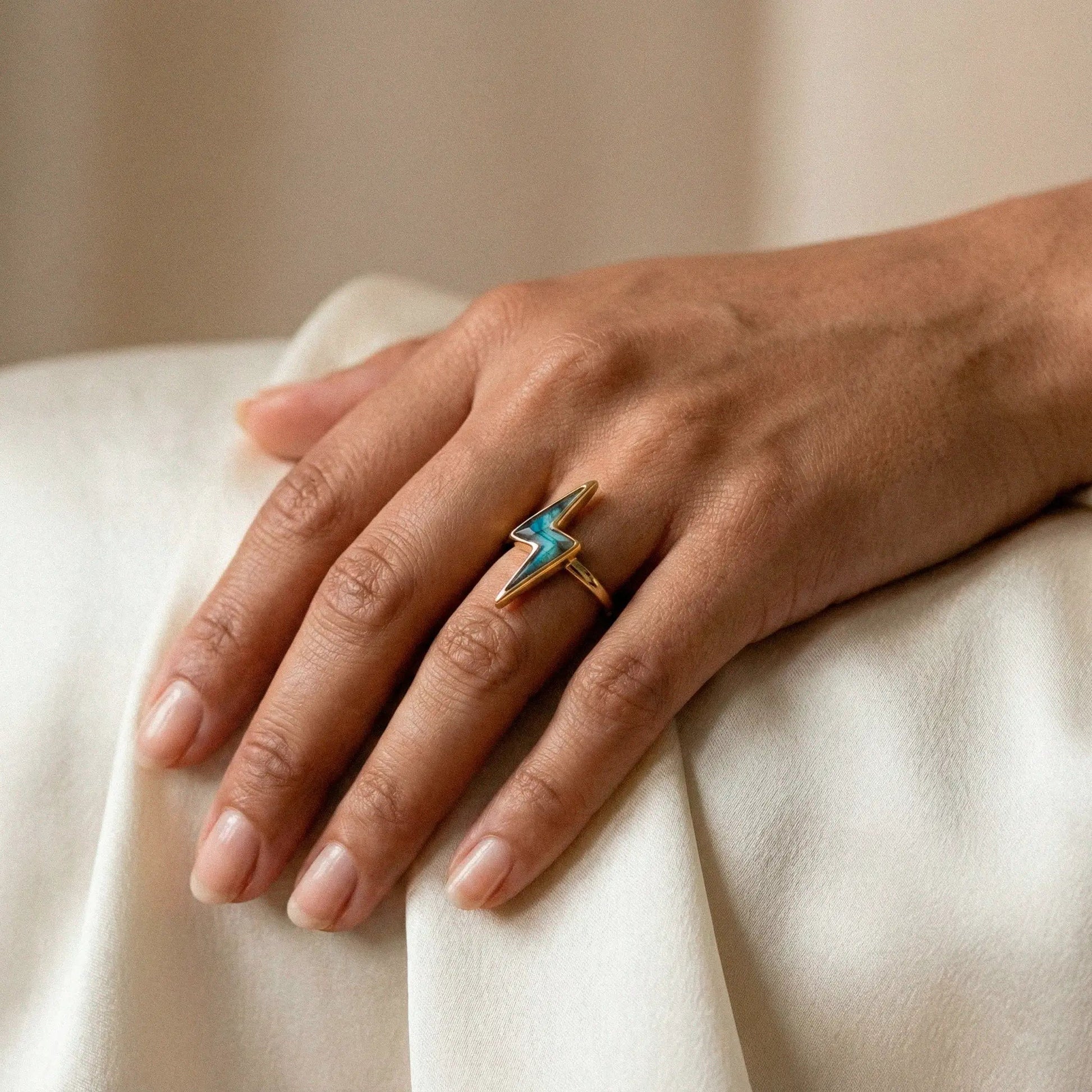 Hand wearing a gold lightning bolt ring with a blue gemstone on a beige background