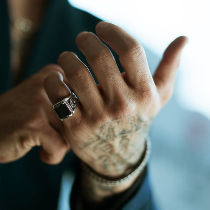 Close-up of a sterling silver laurel ring with an obsidian centerpiece worn on a tattooed hand. The intricate laurel detailing and bold black stone create a sophisticated and powerful statement piece for men.