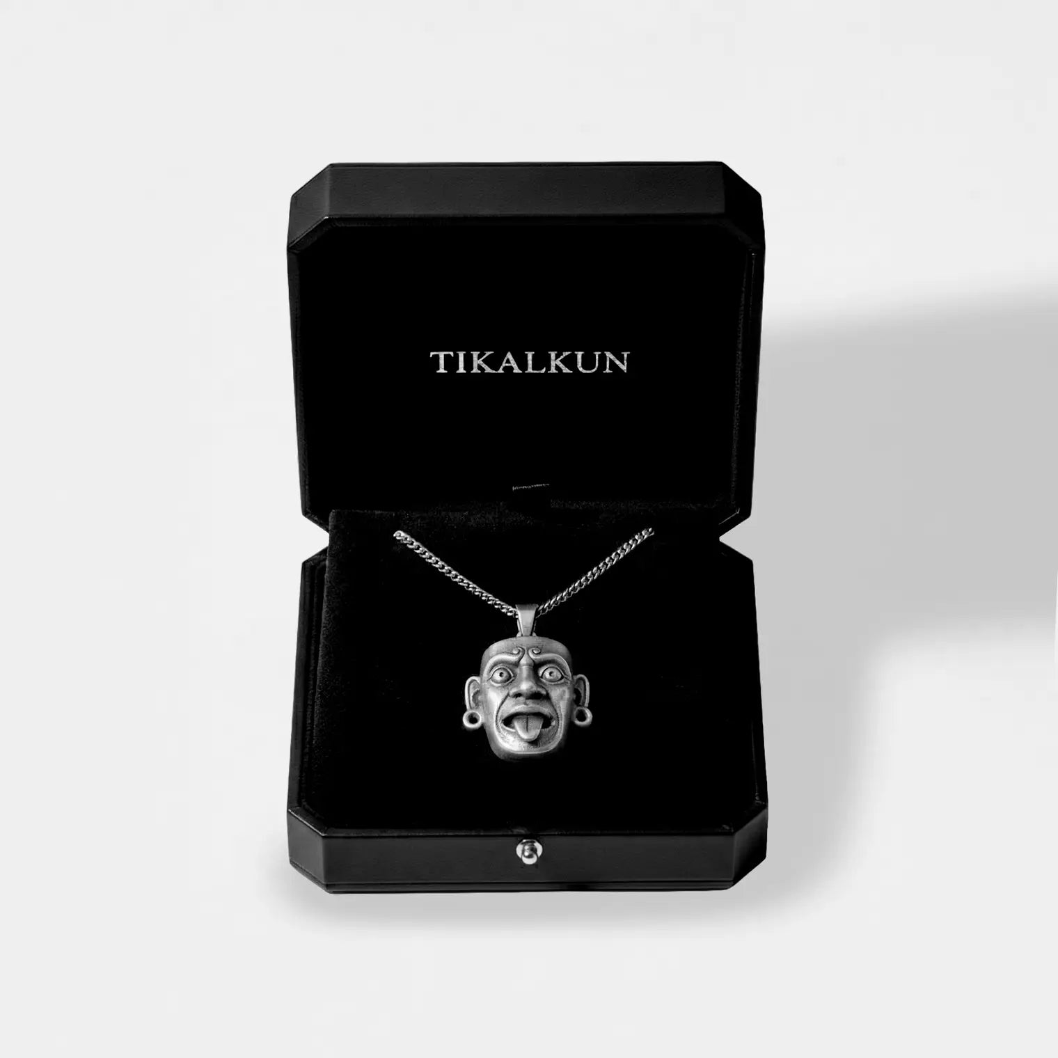 Silver necklace with a mayan mask design in an open black box labeled 'TIKALKUN' on a light gray background.