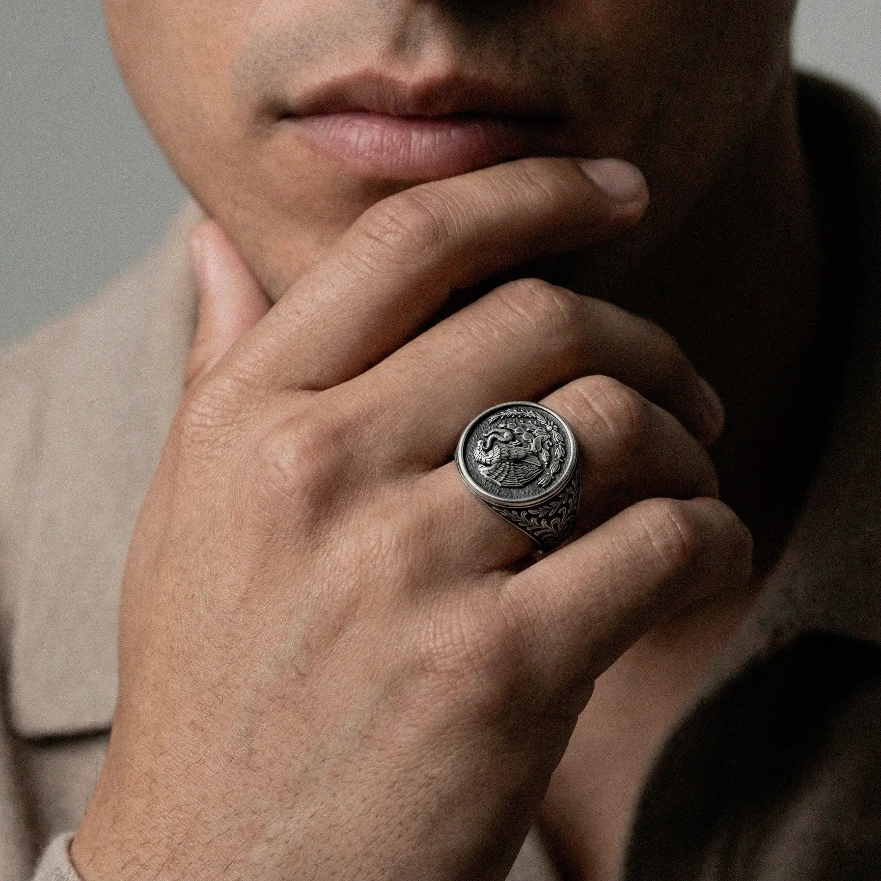 Close-up of a hand wearing a silver ring with a mexico´s national emblem design, holding chin against a neutral background